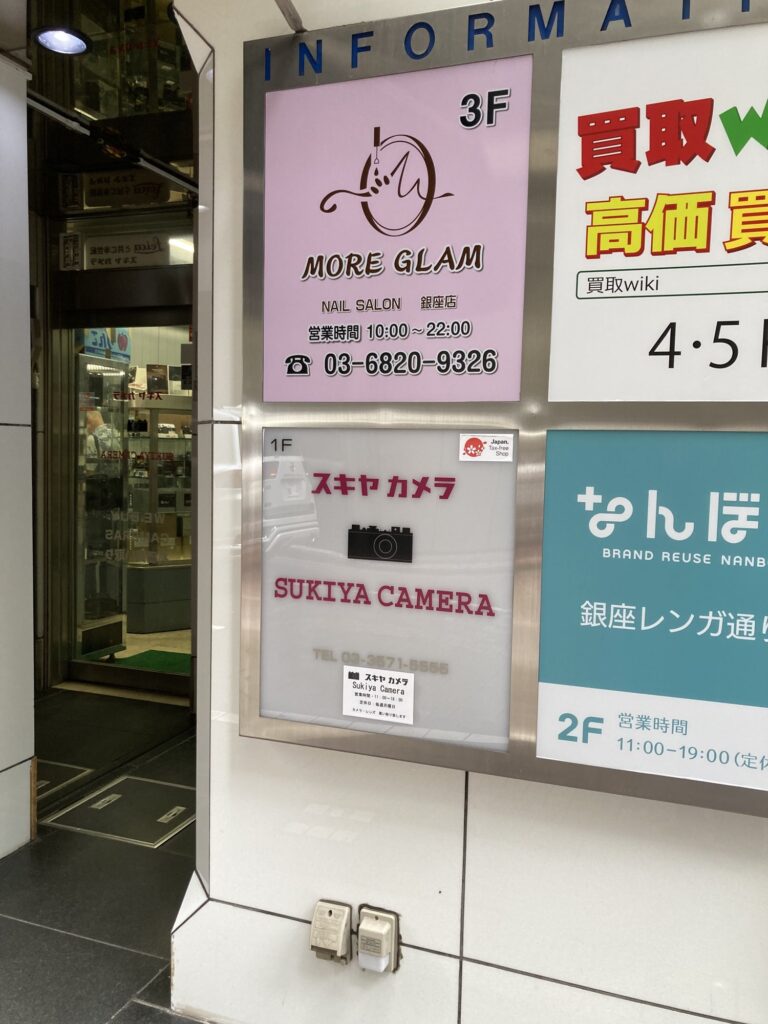Building directory board for Sukiya Camera on the ground floor, with a Japan Tax-free Shop sticker and an opening-hours card